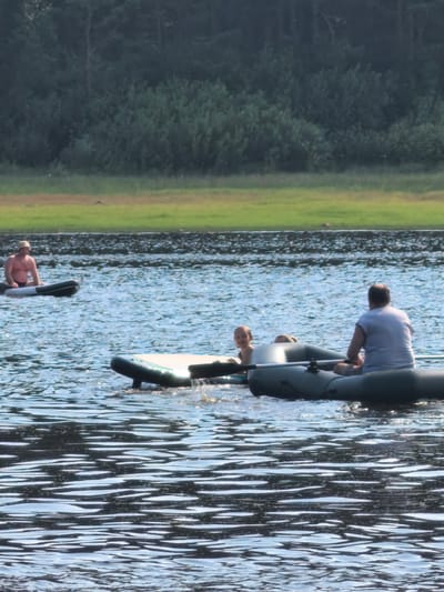 In Sysert, Russia, on a bright, sunny day, three individuals are enjoying recreational activities on a tranquil body of water. In the foreground, a man is seated in a green inflatable boat, facing away from the camera and appearing to be steering. Beside him, a young child with light-colored hair is visible, looking towards the camera. Another inflatable object, possibly a raft or a second boat, is partially submerged in the water in the center foreground, with its dark sides and a light-colored top visible. In the mid-ground, a man is sitting on a black and white inflatable raft, facing left. The background features a dense, dark green forest that stretches across the horizon, meeting a strip of bright green grass and a darker line of rocky shore along the water's edge. The water is a deep blue, with numerous ripples and reflections of sunlight shimmering on its surface.
