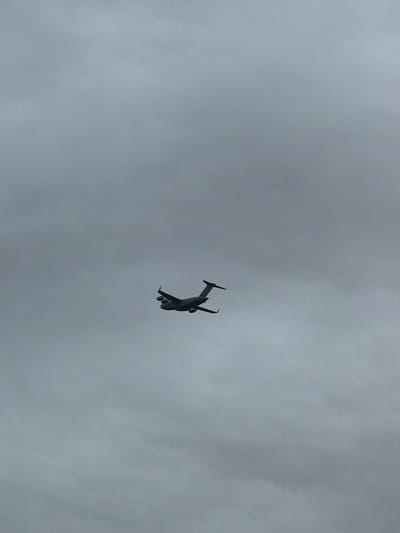 A large, dark gray C-17 Globemaster III military transport aircraft is depicted in flight against a uniformly overcast, light gray sky. The aircraft is observed from below and slightly to its starboard-rear quarter, showcasing its distinctive T-tail and four engine nacelles positioned beneath its high-mounted wings. Its landing gear is fully retracted. The sky is completely covered by dense, layered gray clouds, with no discernible ground features, indicating the aircraft's altitude. No people or other objects are visible. The scene is located in the airspace over Dover, Delaware, United States, consistent with operations near Dover Air Force Base.