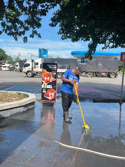 In Ban Ko Pru, Thailand, an elderly man wearing a blue t-shirt, black pants, and grey boots is mopping a wet, tiled ground with a yellow mop. He is standing near a large advertisement sign for KFC, which features pictures of fried chicken meals. In the background, several large trucks are parked under a bright blue sky. A gas station canopy is visible, with blue and white accents. The scene appears to be an outdoor area, possibly a rest stop or a commercial establishment, with trees providing shade in the upper portion of the image. The ground is wet and reflects the surroundings, indicating recent cleaning or rain.