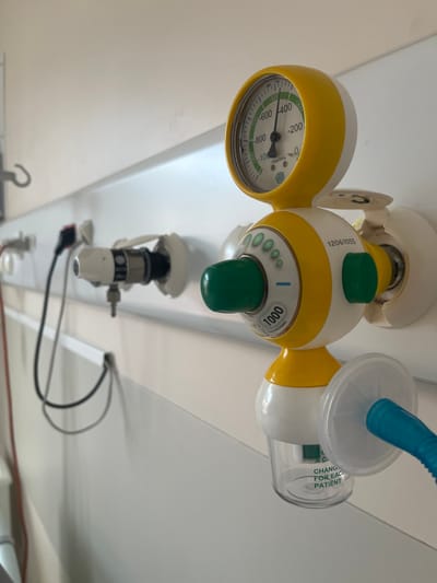 A medical suction regulator unit is depicted mounted on a white wall panel in a healthcare facility within Dumbéa, France. The unit is primarily yellow and white with green control knobs and accents. A circular pressure gauge at the top indicates approximately -700 mbar/hPa on a scale ranging from -1000 to 400 mbar/hPa. Below the gauge, a green rotary knob marked "1000" and a smaller green button are visible, along with the serial number "12061055". A transparent collection jar, bearing the instruction "CHANGE FOR EACH PATIENT", is attached beneath the regulator. This jar connects via a white circular filter to a blue corrugated tube extending rightward out of the frame. To the left, additional medical gas outlets or utility ports are present on the white wall panel, one featuring an attached black cable. A metal hook is mounted further left on the panel. The scene is devoid of human presence or overt activity.