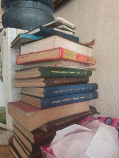 This interior scene, situated in Dogsenang Gorphoog, Bhutan, displays a vertical stack of approximately 16 to 17 aged books of varying sizes and thicknesses. The book covers exhibit diverse colors including red, green, brown, blue, and beige. Several book spines contain visible text in an unidentified script, potentially Dzongkha or Tibetan. A notable red-covered book, approximately midway up the stack, features a yellow and gold decorative spine. Below this, two distinct blue-covered books share a similar gold spine design, which includes a symbol and the inscription "1938-42". In the upper left quadrant, a dark, coarse-textured, partially visible spherical vessel rests upon a white, rectangular object. In the lower right corner, a pink and white package, identifiable as a tissue box by the visible "250" print, contains several crumpled white tissues. The background consists of a plain, light-colored wall. No individuals are present, and no actions are depicted.