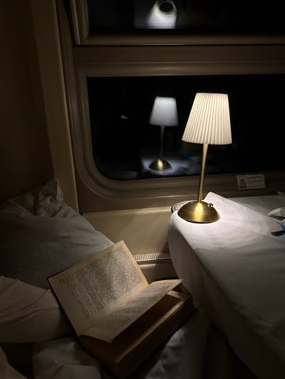 The image depicts the interior of a train compartment at night. A reading lamp with a pleated shade casts light on an open book resting on a pillow-covered seat. The window shows a dark reflection of the lamp. A small informational card is affixed above the lamp. The text on the open book is in Russian. There is no evidence to suggest this is in Lukhovitsy, Russia.