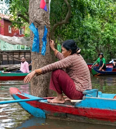 In the city of Soutr Nikom, Cambodia, a woman kneels in a brightly painted blue and red wooden boat, her right hand resting on the rough bark of a large tree. She wears a patterned long-sleeved shirt and dark red trousers. Two pieces of blue plastic are tied to the tree. In the background, a man in a pink shirt sits in another boat, and further back, a person in a green shirt rows a boat. The boats are on murky water, surrounded by lush green trees. Wooden structures and a red house are visible on the left bank.
