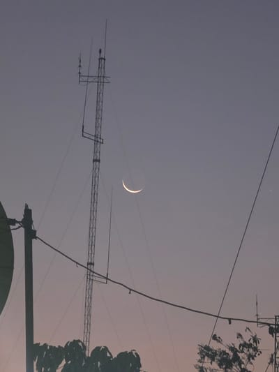 A nighttime scene in Pintomakmur, Indonesia shows a crescent moon in the sky above an urban environment. The sky is a dusky gray. An antenna tower and overhead power lines are silhouetted in the foreground. Plant life is present on the right side of the frame.