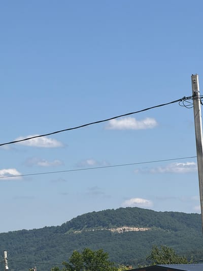 The image is a vertical, medium-shot, eye-level photograph of a natural landscape with man-made elements. The main subjects are a steep, tree-covered mountain slope on the right, and a clear blue sky occupying the left and upper portions of the frame. A power line pole and cables are prominently featured, running horizontally across the upper part of the image and diagonally downwards on the left.
The mountain slope is densely forested with a lighter, rocky strip visible partway up its face. A few white, wispy clouds are scattered across the sky. On the far right, the top of a building with a dark roof and some green foliage are visible, suggesting the edge of a settlement. The image appears to have been taken during the daytime, with bright sunlight indicated by the clear sky and the way light falls on the landscape. The overall impression is one of a rural or mountainous area, possibly in a temperate climate. There is no visible text in the image. The scene is tranquil and depicts a natural environment juxtaposed with utility infrastructure.