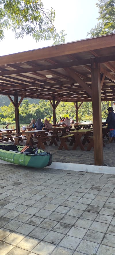 This is an eye-level, outdoor, vertical shot of a covered wooden pavilion with picnic tables in front of a body of water and a wooded hillside. The pavilion has a slatted wooden roof and is supported by wooden posts. Several groups of people are seated at the picnic tables, some appearing to be eating or socializing. The ground within the pavilion and extending outwards is paved with square tiles. To the left of the pavilion, on the tiled ground, rests an inflatable kayak that is green and white. The water appears calm, and the surrounding vegetation is lush and green. The sky is bright and clear, suggesting a sunny day. This scene is located at the Aqua Marina water sports facility in Lamsenia, Aceh, Indonesia.