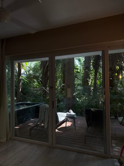 The image depicts an interior view looking out through sliding glass doors onto an outdoor patio and lush tropical vegetation, located in Punta del Burro, Mexico.

The foreground shows the interior of a room with a light-colored wooden or laminate floor and a light brown ceiling. A white ceiling fan is partially visible in the upper left corner. A light-colored curtain is drawn to the far left. The white-framed sliding glass doors occupy the central and lower portions of the image.

Beyond the glass doors, an outdoor wooden deck is visible. On the left side of the deck, a rectangular hot tub or small plunge pool with dark tiled sides (appearing blue-black) contains water. A silver-colored handrail is mounted on the edge of the tub. A light-colored towel is draped over a structure near the hot tub. In the center-right of the deck, a light-colored lounge chair with a striped cushion is present, facing away from the viewer. A small, dark side table next to this lounge chair holds a white rolled towel. To the far right, a dark-colored lounge chair is partially visible.

The area beyond the deck is characterized by dense, vibrant tropical foliage, including numerous tall palm trees and various broad-leafed plants. Sunlight filters through the canopy, illuminating parts of the vegetation and casting shadows on the deck. No individuals or active subjects are present in the scene.