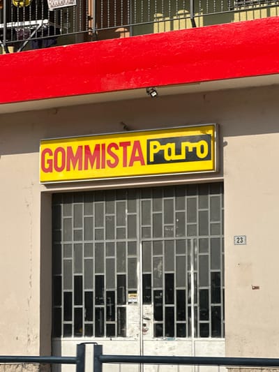 A vertical, rectangular yellow sign with red lettering hangs on a beige building wall. The sign reads "GOMMISTA Pa.ro". Next to the sign is a window with a grey metal security grate. Above the window, a small blue and white number "23" is displayed on the wall. To the left of the sign and window, a bright red, flat surface is visible, likely a painted wall or door. In the background, to the left of the red surface, metal railings and a chain-link fence suggest an outdoor area. The lighting and shadows indicate it is daytime, likely sunny. The scene appears to be a storefront or business in Rudiano, Italy, given the context. There are no people visible in the image.