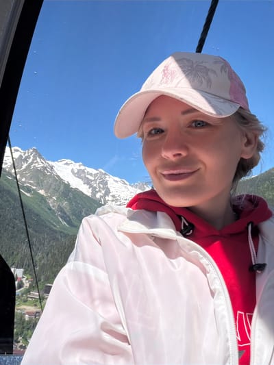 A woman is taking a selfie inside a cable car with snow-capped mountains in the background. She is wearing a pink cap with a palm tree pattern, a red hooded sweatshirt, and a white jacket. The cable car appears to be moving, as evidenced by the slight blur in the background. The sky is clear and blue. The scene is set in Dombay, Russia, a mountain resort town known for its skiing and hiking.