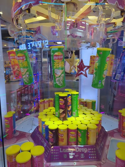 This image depicts an indoor scene, likely a game arcade or entertainment center, in the Xishan District of China. The primary focus is a "Lucky Drop" claw machine filled with cylindrical snack containers, predominantly Lay's potato chips. The machine features a clear glass front, revealing the prizes within and the mechanical claw apparatus suspended above. The snacks are arranged in multiple rows, with colorful packaging in shades of red, green, and yellow. Some containers are hanging, suggesting they are prizes waiting to be claimed, while others are stacked on a tiered platform at the bottom. The machine itself is adorned with bright, reflective lighting and decorative elements like stars. The backdrop shows a blurred view of a commercial establishment, with more arcade machines and display lights visible through the glass. There are no people clearly visible interacting with the machine.