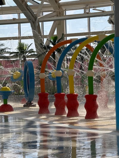 The image depicts an indoor water park installation in Kogalym, Russia. The structure consists of a series of brightly colored, curved pipes (green, yellow, blue, orange, and blue) connected to red nozzles. Water is actively spraying from the nozzles. The pipes and nozzles are mounted on a wall, where water is flowing down. Through the glass roof, palm trees and the sky are visible in the background. Structural steel beams support the roof.