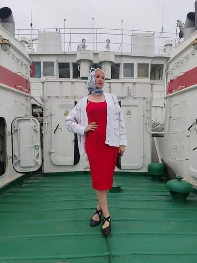 A woman with pink hair, wearing a red dress, a white cardigan with heart patterns, and a blue and white patterned headscarf, stands on the deck of a ferry boat. She has her left hand on her hip and is looking to her left. The ferry is white with red stripes and has multiple doors and hatches on its sides. The deck is painted green. In the background, more parts of the ferry are visible, including cabins with windows and railings. Two green bollards are present on the deck. The sky is overcast, suggesting it is daytime. The scene appears to be a candid photo taken on a ferry, possibly in Bereg, Russia, given the location context. There is no visible text that can be read clearly in the image.