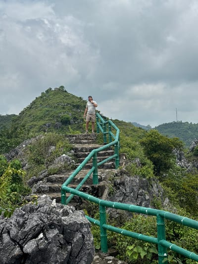 A man stands on a stone staircase on a rocky, vegetated mountainside in Tam Thanh Ward, Vietnam. The man, wearing a white t-shirt, tan shorts, and a dark beanie, is looking towards the camera and holding his hand to his mouth. A green railing lines the staircase, which ascends a steep incline. Lush green vegetation covers the rocky terrain, with a prominent, tree-covered peak in the background. The sky above is overcast with heavy clouds.