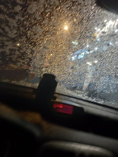 A blurry view through a car windshield covered in rain or snow, illuminated by streetlights and traffic. A dashboard camera is mounted to the windshield, its red light visible. The interior of the car is dark, with a portion of the dashboard and possibly a seat visible on the right. The overall impression is of driving in inclement weather at night.