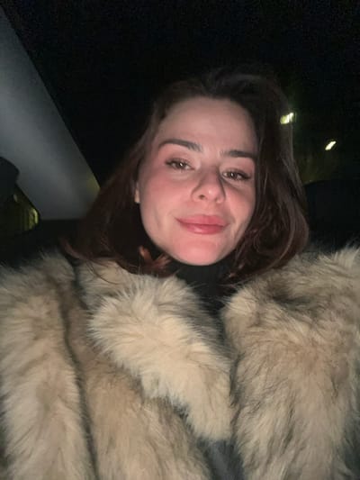 A fair-skinned woman with dark auburn hair, wearing a light brown fur coat, poses closely for a selfie. Her dark eyes, eyebrows, and lips are prominent. The lighting appears to be low, creating shadows on her face and background. The texture of the fur coat is detailed, contrasting with the dark, blurred space behind her. The image orientation is tilted slightly.