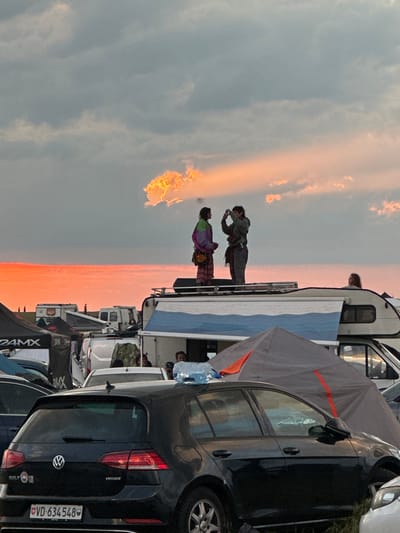 The image shows a sunset scene at what appears to be a campground or outdoor event, possibly in Bengy-sur-Craon, France. The sky is divided between gray clouds and a bright orange and pink sunset. In the foreground, various vehicles are parked, including a black Volkswagen Golf with the license plate "VD-634548." A tent or awning is partially visible near the car. Further back, people are standing on the roof of a trailer or RV, seemingly taking pictures of the sunset. The setting suggests an outdoor event, possibly a festival or gathering, in the rural or suburban area of Bengy-sur-Craon.