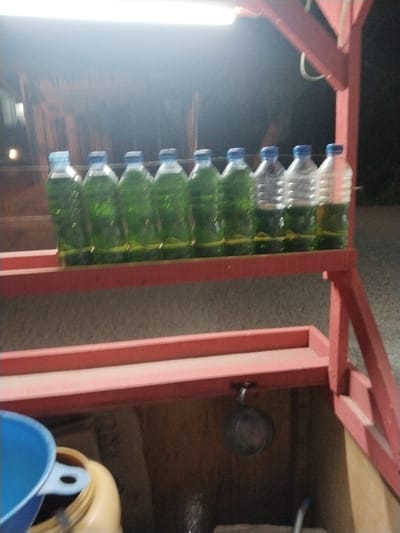 The image displays a nighttime scene, identified as Aceh Harbor, Indonesia. In the foreground, a red-painted display stand, likely constructed of wood or metal, is brightly illuminated by an overhead rectangular light fixture. The stand features at least two shelves. The upper shelf holds nine uniform plastic bottles. Seven bottles, positioned from the left, contain a translucent green liquid and are sealed with blue caps. The two bottles on the far right contain a translucent yellowish liquid and are also sealed with blue caps. The lower shelf of the stand is empty. Within the base compartment of the stand, a blue plastic funnel is visible on the left, adjacent to a light-colored plastic container holding a dark liquid. A small metal pan or scoop hangs from the underside of the upper shelf. The background is dark and out of focus, showing indistinct structural forms, potentially buildings, under the ambient light. No individuals, actions, or direct interactions are depicted.