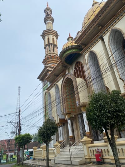This image captures a wide-angle view of a large, ornate mosque situated along a road in Jepara, Indonesia, under an overcast sky. The mosque, constructed primarily with light cream-colored walls and dark marble-like accents, features a prominent, multi-tiered minaret on its left, decorated with intricate geometric patterns in beige, brown, and gold, topped with a pointed finial. Two loudspeakers are visible on the minaret. A large, golden-yellow main dome with ornate banding and a finial dominates the mosque's roofline, accompanied by a smaller, similar dome. Below the large dome, a section is adorned with green and dark blue trim featuring white Arabic calligraphy.
The mosque's facade includes multiple large, arched windows and recessed sections with patterned tiling in shades of green, gold, and white. Ornate columns with golden capitals and bases support architectural elements. A wide, light-colored staircase with stainless steel railings leads up to an elevated entrance, bordered by a balustrade. On the ground near the stairs, a red cylindrical bin, possibly a waste receptacle or fire safety cabinet, displays a white symbol (appears to be a "No Smoking" sign).
Along the tiled sidewalk in front of the mosque, three small, leafy trees with slender trunks are planted. Dense green bushes are visible near the mosque's lower facade. In the background on the left, an asphalt road runs parallel to the mosque. Multiple utility poles with a dense network of black electrical and communication wires crisscross the sky, extending across the frame. Further in the distance, a tall, metallic lattice communications tower stands against the grey sky. Beyond the road, glimpses of residential or commercial buildings with reddish-brown tiled roofs are visible. On the far left, in the background, two indistinct figures are visible near a parked scooter or motorbike, one appearing to be seated on it.