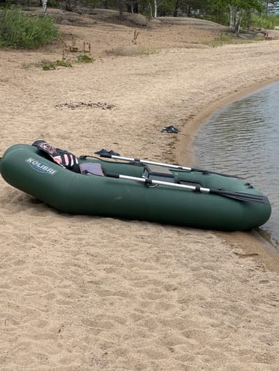 An inflatable, dark green Kolibri brand boat lies partially on a sandy shore and partially in calm, rippling water. The boat is oriented lengthwise, with its bow pointing towards the water. Two oars, with black blades and silver shafts, are secured to the boat and rest on top of it. Near the center of the boat, a black and pink life vest or seat cushion is visible. The boat has a visible logo that reads "KOLIBRI" in white text with a blue outline, and below it, "WWW.KOLIBRI.UA".

The scene is a beach with light-colored sand that shows some footprints and small debris. The water's edge is defined by a gentle curve in the shoreline. In the background, to the left, there is a wooded area with trees, including some birch trees with white bark, and rocky formations. The lighting suggests it is daytime, and the weather appears to be overcast or cloudy, as there are no strong shadows. The overall impression is a peaceful, outdoor setting, likely a lake or riverbank in Aiyrtau, Kazakhstan.