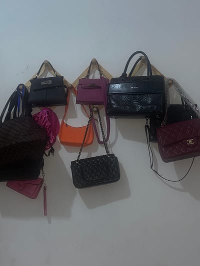 A wall-mounted wooden rack displays an assortment of handbags. The bags vary in color and style, including black, purple, orange, pink, and burgundy. Some of the bags are quilted, while others have smooth textures. The scene is indoors against a plain white wall. The location is not specified as being in Abraka, Nigeria.