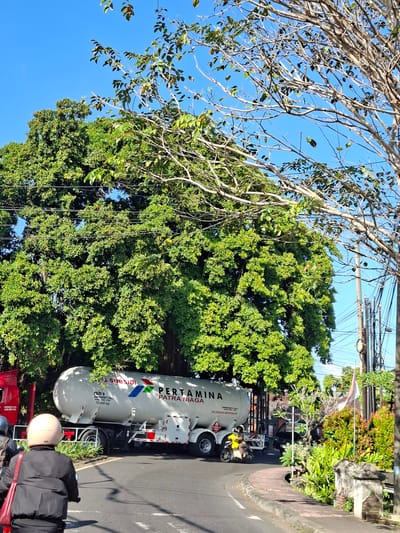 In Banjarangkan, Indonesia, on a clear, sunny day, a large white Pertamina Patra Niaga tanker truck is parked on a paved road, partially obscured by a dense green tree. The truck's tank is marked with the Pertamina logo and text, including "SUBSIDI," "EMERGENCY CALL," and "DILARANG MEROKOK" (meaning "No Smoking"). A red truck is partially visible to the left of the tanker. In the foreground, two people on motorcycles are riding away from the camera. The person in front is wearing a black jacket and a light-colored helmet, with a red bag slung over their shoulder. The person behind them is wearing a grey helmet. The road curves to the right, with a sidewalk and lush vegetation bordering it. Power lines and poles are visible in the background, along with a national flag and more trees.