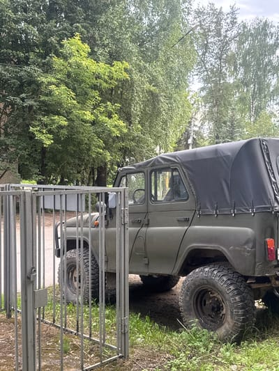 A parked, olive-drab military-style SUV with a black canvas roof is visible in front of a metal gate. The vehicle has large, rugged tires and an exposed spare tire on the rear. The gate is made of vertical metal bars and appears to be closed. The background consists of lush green trees and foliage, with a hint of a building on the left side. The ground is covered with dirt and grass. The scene appears to be in an outdoor urban or suburban setting, possibly a yard or parking area. The image was taken in Dedovsk, Russia. There are no people visible in the image.