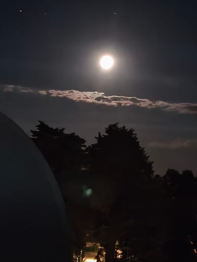 The image depicts a nighttime scene in Passignano sul Trasimeno, Italy, under a full moon. The moon is the dominant light source, appearing as a bright, luminous orb in the dark sky. Thin, wispy clouds are illuminated by the moonlight, creating a soft glow around them. Below the sky, the silhouettes of trees are visible, their branches stark against the night sky. A large, dark, curved object occupies the left side of the foreground, partially obscuring the view of the trees. This object appears to be artificial, possibly a building or structure. The lower portion of the image shows faint lights, suggesting the presence of artificial illumination in the distance, indicative of the town. There are no visible people or animals in the frame.