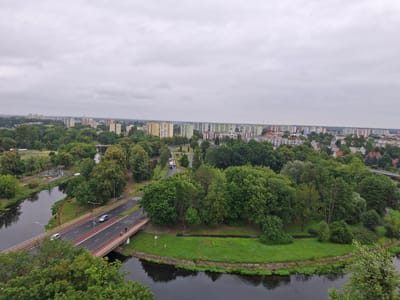 This is an aerial, wide-angle photograph capturing a cityscape under an overcast sky. The scene depicts a river flowing through a densely vegetated urban area, with a bridge crossing the water. Several cars are visible on the road on the bridge, and a few people are seen walking on the sidewalk.
In the background, a sprawling collection of apartment buildings, mostly in muted colors with some featuring yellow or green facades, dominates the skyline. These buildings are interspersed with trees, suggesting a city with a significant amount of green space. The overall impression is one of a developed urban environment integrated with nature. The weather appears to be cool and perhaps humid, given the grey sky and lush greenery. The time of day is indeterminate due to the overcast conditions. The image provides a sense of the layout of the city, with residential areas, a river, and a bridge connecting different parts of the landscape. No visible text is present in the image.