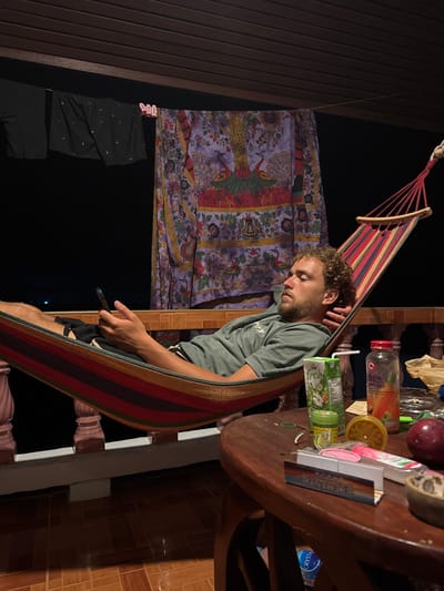 The image depicts an outdoor night scene on a tiled balcony in Haad Yao, Thailand. A light-skinned male, with curly brown hair and a beard, is resting in a striped hammock. He is wearing a greenish-gray t-shirt and dark shorts. His left hand holds a dark smartphone, and his eyes are closed or partially closed. The hammock features prominent red, orange, yellow, green, and blue stripes.
Behind the hammock, a clothesline is strung across the balcony, from which two dark garments and a large purple tapestry adorned with peacock and floral patterns are hanging. The background beyond the balcony is dark, indicative of nighttime, with faint distant lights visible. The balcony itself features a wooden railing with light-colored balusters.
In the foreground, to the right, a dark brown oval wooden table is laden with several objects: a green carton of "fresh" juice with a straw, a clear bottle labeled "100% CARROT JUICE," a small green container, a yellow, possibly dried citrus rind, a red apple, a small bowl containing dark material, a crumpled light-colored bag, and a rectangular packet of rolling papers. The balcony floor is covered in brown tiles.