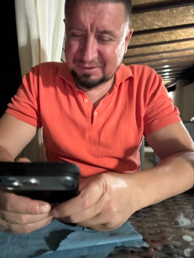 A man wearing an orange polo shirt sits at a table outdoors, possibly on a patio, as suggested by the wooden ceiling structure and translucent curtains in the background. He is looking down at a black object, likely a smartphone, held in his hands. The man's face shows signs of distress or discomfort, with furrowed brows and his eyes closed. A light blue cloth is visible beneath the object he is holding. The overall lighting suggests it is evening or nighttime. The setting appears to be within a restaurant or cafe environment in Kurortne, Ukraine.