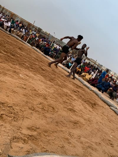 The image shows a wrestling match taking place on a sandy field in Sokoto, Nigeria. Two shirtless men wearing shorts are wrestling on the field. The man on the left is attempting to throw the man on the right to the ground. The men are surrounded by a crowd of spectators sitting along the edge of the field on sandbags. The wrestling match is taking place in an open area, most likely an outdoor arena or training field. The spectators are wearing a variety of clothing, including traditional Nigerian garb. The environment is dusty, indicating that it is likely a dry season. The sandy field is bare, with only a few footprints and the sandbags marking the perimeter of the wrestling area. The sky is clear and the weather is sunny. There are a few buildings in the background, and the atmosphere is energetic and lively.