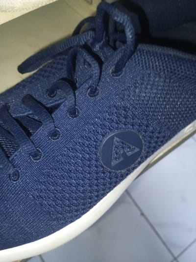 A close-up shows a navy blue sneaker with a white sole. The shoe has a textured knit upper, and the laces are also navy blue. A small, circular logo featuring a triangle with a stylized 'A' inside is visible on the side of the shoe. The image is well-lit.