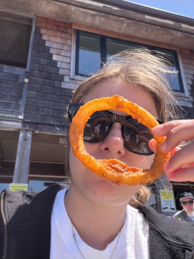A young person wearing sunglasses and a black jacket over a white t-shirt is holding a large, battered onion ring up to their face, obscuring their eyes. The onion ring is golden brown and appears to be fried. The background shows the exterior of a wooden building with shingled siding and dark windows, likely a restaurant or shop, under a bright blue sky. In the distance, another person wearing a baseball cap and sunglasses is visible, as well as a yellow sign that says "CUSTOMER PARKING". The scene is set in Cannon Beach, Oregon, United States of America, a coastal town known for its scenic beaches and arts scene.