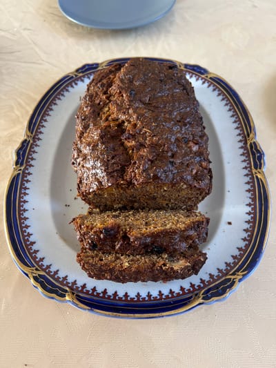A loaf of dark brown, moist-looking baked goods, possibly bread or cake, sits on a decorative oval platter. The platter is white with a blue and gold patterned rim. Three slices have been cut from the left side of the loaf. The baked good appears to contain visible chunks of nuts and possibly dried fruit. The platter is placed on a light-colored, textured tablecloth. In the upper portion of the image, a portion of a light blue plate is visible. The image was taken from a high-angle perspective. The location is not specified in the provided data.