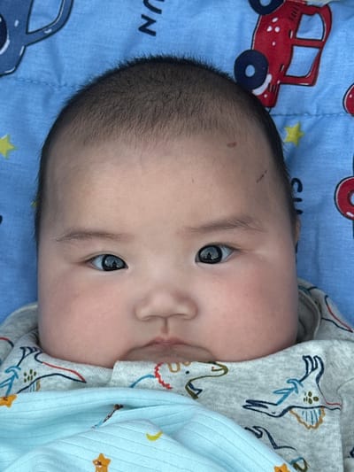 The image is a close-up, rotated photograph of a baby lying on a blue blanket with car and dinosaur motifs. The baby's face is the primary subject, with a portion of their head and upper body visible. The baby has dark hair and appears to be very young. Their right eye is open and clearly visible, reflecting light. The baby is wearing a grey onesie with a pattern of blue and brown dinosaurs, and is partially covered by a light blue blanket.

The blue blanket has a repeating pattern of red cars, yellow stars, and what appear to be moons and other celestial shapes. The word "UN" is partially visible in black text on the blanket near the baby's head.

The lighting suggests it is daytime. There are no explicit cues about the weather or specific location beyond what can be inferred from the baby's clothing and the blanket's pattern. The baby's expression is neutral or contemplative, with no strong emotional display. The overall scene is intimate and personal, focusing on the infant.