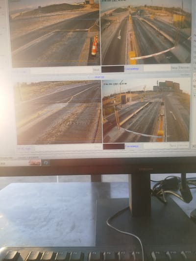 The image shows a computer monitor displaying a surveillance system with four live camera feeds. The feeds show a road or checkpoint with a boom gate and traffic cones. The timestamps indicate the date as June 27, 2026, and the time as 06:03:00. The location is identified as "Түндүк айланып өтүүчү жол 84 га, Kyrgyzstan," which translates to "Northern Bypass Road 84 ha, Kyrgyzstan." The text on the screen also includes labels like "Госномер" (License Plate) and "Ввод Расп." (Entry/Exit). In the foreground, a keyboard is visible. The overall scene suggests a traffic monitoring or control point.