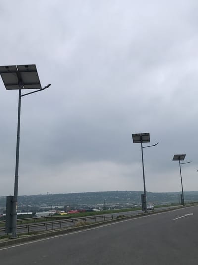The image depicts a daytime scene in Șcheia, Romania, under an overcast, light grey sky. A multi-lane asphalt road, sloping slightly uphill to the right, occupies the foreground and midground, featuring white arrow road markings on the right lane. Metal guardrails line the road's edge. Three grey solar-powered streetlights are visible on the right side of the road, each comprising a tall pole, two rectangular solar panels mounted atop, and an LED light fixture. A fourth streetlight is partially visible on the far left. In the mid-distance, a light-colored car travels on the road. The background reveals a sprawling cityscape or town built into rolling hills, characterized by numerous buildings, some with industrial or commercial appearances (e.g., red and yellow roofs), and residential structures. A prominent water tower stands on a distant hill, serving as a landmark. Sparse vegetation and grass are present along the roadside and throughout the distant landscape.