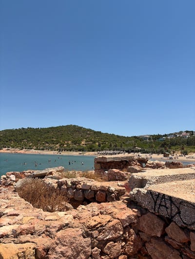 The image depicts a coastal scene in Vouliagmeni Municipal Unit, Greece, on a clear, sunny day. In the foreground, there are weathered, reddish-brown stone ruins with visible construction materials and some graffiti. Beyond the ruins, a body of tranquil, turquoise water is populated with numerous individuals swimming and enjoying the sea. The shoreline features a sandy beach dotted with sun umbrellas, beachgoers, and some vegetation. Further inland, a verdant, tree-covered hill rises, with buildings visible on its slopes and at its base, suggesting a populated area. The sky is a uniform, bright blue with no clouds.