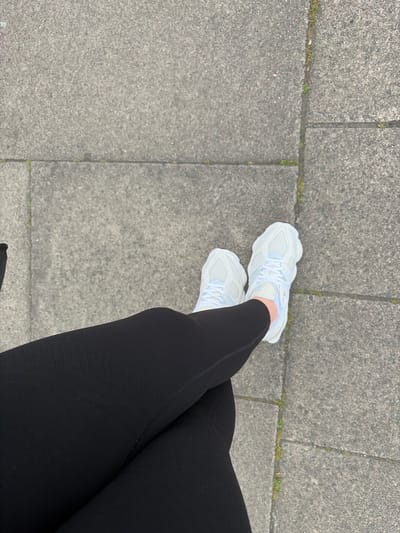 The image shows a person's lower body and legs standing on a gray concrete pavement. The person is wearing black leggings and white sneakers. The pavement is made up of square slabs, and there are small patches of green moss growing in the cracks between the slabs. The exact location in Leicester, United Kingdom, cannot be determined from this image alone.