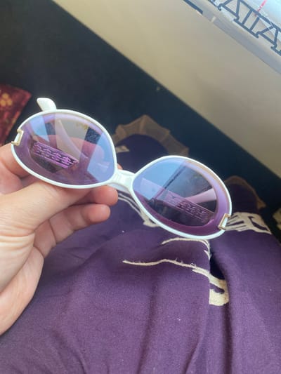 The image shows a pair of white and purple Chanel sunglasses being held by a hand. The sunglasses have purple lenses and a white frame with gold accents. The background is a dark purple fabric with a white pattern. There is no indication of the location being Arbagish, Uzbekistan. The image appears to be taken indoors or in a shaded outdoor area.