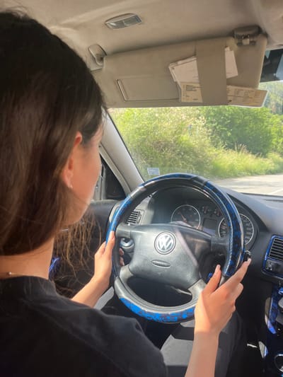 A person with long, dark brown hair is driving a car. The viewpoint is from behind the driver, looking forward. The driver is wearing a black t-shirt and has her hands on a steering wheel with a blue and black pattern. The car's dashboard and steering wheel are visible. The car appears to be moving, as indicated by the blurred background of greenery and a road. The sun is shining, casting shadows inside the car. The car's sun visor is down and has papers clipped to it. There are no other people visible in the car. The environment outside the car suggests a rural or semi-rural setting, with lush green trees and vegetation. The image appears to have been taken on a road in or near Vlahovo, Bulgaria, based on the provided context.