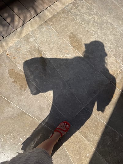 The image shows a section of tiled flooring with a large shadow cast upon it. The shadow appears to be of a person. A leg and foot wearing a red sandal are visible on the right side of the image, partially obscured by the shadow. The lower portion of a gray, textured garment is seen on the bottom right corner. The tiles are light gray and beige in color. This image was taken in the city of El Gouna, Egypt.