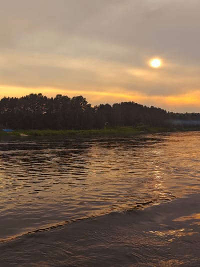 A photograph captures a sunset over a wide river, likely the Kama River, near the village of Yukseevo, Russia. The sky is a muted orange and yellow, with thin clouds partially obscuring the sun, which appears as a bright, hazy orb in the upper right quadrant of the image. The foreground is dominated by the river's surface, reflecting the warm hues of the sky and displaying ripples and a small wave generated by movement, possibly from a boat. On the far bank, a dense line of dark trees forms a silhouetted treeline against the horizon. Some green vegetation is visible at the base of the treeline. A faint plume of smoke rises from the treeline in the middle distance on the right side of the image. On the left bank, there are a couple of small, blue objects, possibly boats or tarpaulins, and a patch of light-colored sand. The overall impression is one of serene, natural beauty at dusk.