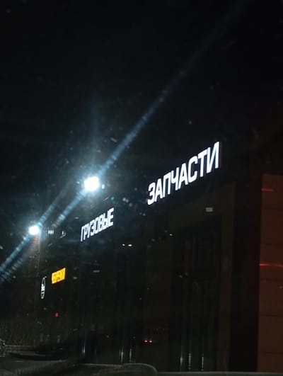 The image shows a nighttime scene of a building in Enem, Russia. The building has two lit signs: "ГРУЗОВЫЕ" (Freight) and "ЗАПЧАСТИ" (Spare parts). The lights on the signs and streetlights are visible. The overall environment appears dark, with a slight glare on the camera lens. The location is likely a shop or warehouse specializing in spare parts for freight vehicles.