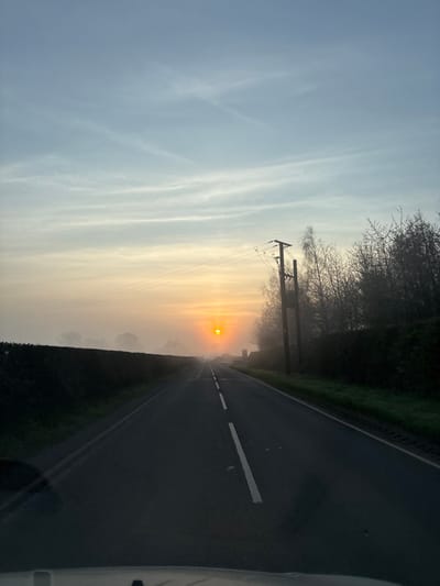 The image displays a straight, dark grey asphalt road in Baschurch, United Kingdom, extending into the distance under a misty sunrise. A bright, intense orange-yellow sun is visible low on the horizon, partially diffused by widespread fog or mist that covers the lower and middle ground. The sky above is light blue, transitioning to pale yellow near the horizon, with subtle wispy cloud formations. Dark green hedges line both sides of the road. On the right, a wooden utility pole with multiple horizontal wires stands upright beside the road, backed by a cluster of bare-branched deciduous trees and shrubs. The perspective suggests the photograph was taken from inside a vehicle, with the dark upper edge of a dashboard or hood partially visible at the bottom of the frame. No individuals are present.