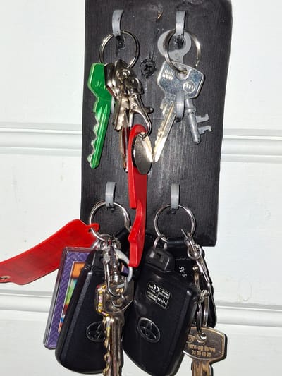 A key holder made of dark wood is mounted on a white wall in Åsgårdstrand, Norway. The key holder has multiple hooks, each holding one or more keys.
The keys are of various types and colors, including silver, green, and red. Some of the keys are attached to keychains, one of which has a red strap. Two of the keys are black car key fobs with a Toyota logo. One of the key fobs has a sticker with text including "MOROCCO" and "APPROVED." Another key has text that appears to be "jern og brøve" followed by numbers.
The scene is well-lit, and the details of the keys and key holder are clearly visible. The background is a white wall with some subtle texture.