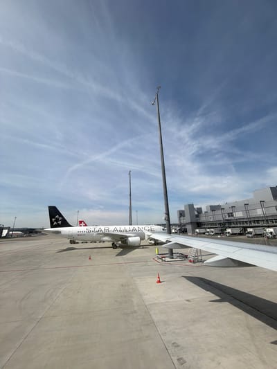 This landscape-oriented image captures a bright, daytime scene at an airport apron in Kloten, Switzerland. The central subject is a large, white passenger airplane, emblazoned with "STAR ALLIANCE" text along its fuselage. Its registration number, HB-UJO, is visible near the tail. A red and white Swiss flag emblem is also prominent on the aircraft's body, and the black tailfin features distinctive white, star-like logos. One of the aircraft's wings extends towards the right, casting a clear shadow on the concrete ground.
To the left of the aircraft, a tall, slender light pole or antenna reaches high into the sky, with another shorter pole visible further left. In the background, a multi-story airport building, likely a terminal or maintenance facility, is depicted with various parked ground support vehicles and equipment. The concrete tarmac is marked with lines and features an orange traffic cone near the aircraft.
The sky is a clear, light blue, with scattered, wispy white clouds indicating fair weather conditions. The bright daylight and the angle of the shadows suggest it is mid-day or afternoon. The scene conveys an impression of an active airport environment, with the aircraft positioned for ground operations or awaiting departure or arrival. No specific human figures are clearly visible, but the overall setup indicates ongoing airport activity.