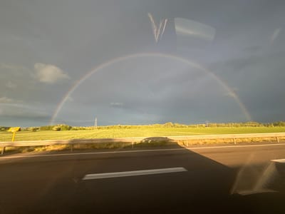 The image depicts a scene from a vehicle on a road, possibly in Orschwiller, France, given the context. A partial rainbow is visible in the sky, arching over a landscape featuring a green field bordering the road. The road itself is asphalt with white lane markings. There is a metal safety barrier running along the roadside. A power line support structure is seen in the distance. Reflections on the vehicle's window obscure the view partially, revealing part of the vehicle's interior.
