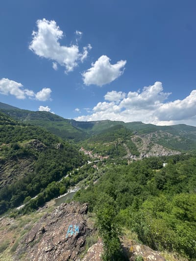 The image captures a panoramic view of a lush, mountainous valley in Bov, Bulgaria, under a clear blue sky dotted with scattered white cumulus clouds. The scene is dominated by steep, forested mountainsides covered in dense green deciduous and coniferous trees. In the mid-ground, a valley cuts through the landscape, revealing a winding river with greenish-brown water, flanked by a narrow paved road. A small settlement with multiple buildings featuring orange or red roofs is visible along the riverbank on the left side of the valley. A white bridge spans the river to the right of the village, connecting to the road. The immediate foreground features a large, dark, reddish-brown rocky outcrop with visible lighter mineral deposits and some sparse green vegetation. Blue graffiti, forming characters resembling "GH," is present on the surface of this foreground rock. No people or animals are visible within the frame. The lighting suggests a bright, sunny daytime environment.
