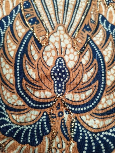 The image shows a close-up of fabric with a symmetrical design. It features an abstract floral or bird-like pattern in shades of brown, white, and dark blue. Small, regularly spaced dots form outlines and fill certain areas. A shimmering golden thread accentuates the central axis of the design. The texture of the fabric is visible, with a slightly coarse weave. The composition is primarily focused on the central motif, showcasing the details of the pattern.